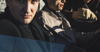 Dwóch mężczyzn siedzących w samochodzie, jeden z nich patrzy w obiektyw, drugi przed siebie. W tle mężczyzna zaglądający do samochodu przez otwartą szybę.