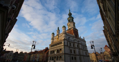 Zdjęcie budynku Ratusza w stylu renesansowym, z jedną wieżą i trzema małymi wieżyczkami na tle biało-błękitnego nieba. Za i z prawej strony Ratusza zaytkowe kamieniczki.