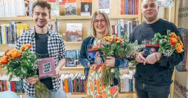 Dwóch mężczyzn i jedna kobieta stoją uśmiechnięci z bukietami kwiatów i statuetkami na tle półek z książkami.