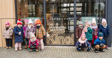 Grupa dzieci stojących na tle szklanej ściany. Pomiędzy dziećmi napis "Ale Kino!".