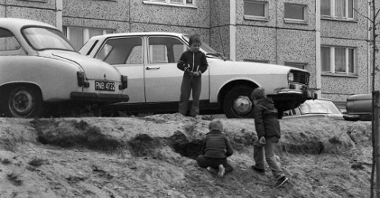 Czarno-białe zdjęcie trójki dzieci bawiących się na poboczu osiedlowej drogi. Za nimi dwa zaparkowane samochody, a w tle blok mieszkalny.