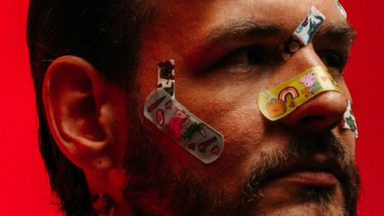 Zdjęcie twarzy mężczyzny z naklejonymi na nosie, policzkach i czole kolorowymi plasterkami. Czerwone tło.