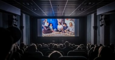 Widzowie w kinie oglądają film na dużym ekranie. Jest ciemno, salę rozświetla tylko światło z ekranu.