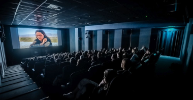 Zaciemnione kino, trwa seans filmowy. Widzów siedzących w fotelach oświetla tylko światło z ekranu.