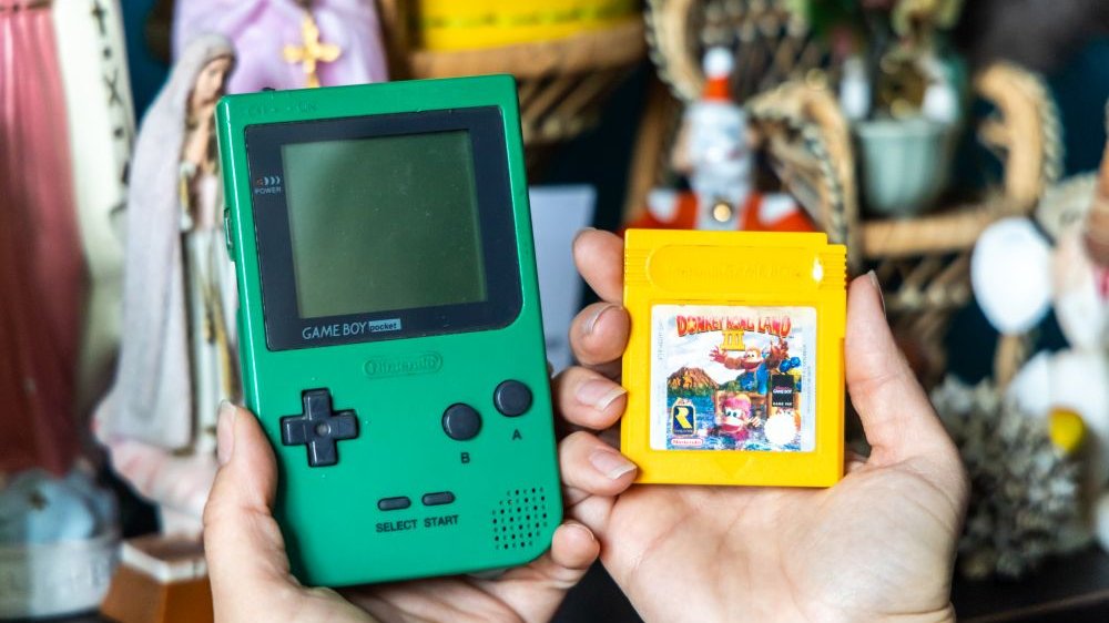 Mini konsole do gier, w tym Game Boy, trzymane w dłoniach. W tle różne przedmioty, na zdjęciu niesotre, w tym figurka Matki Boskiej.