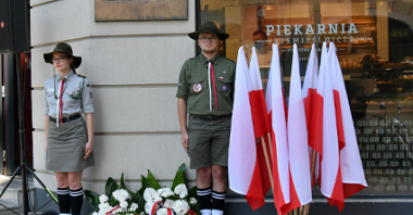 Tablica pamięci Romka Strzałkowskiego, obok harcerze i polskie flagi, pod tablicą kwiaty