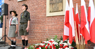 Zdjęcie tablicy pamiątkowej na scianie szpitala. Pod nią kwiaty, obok hacerze i polskie flagi