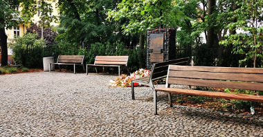 Skwer Trzech Tramwajrek. Na zdjęciu widać ławki, za nimi stoi pomnik, w tle wiele drzew. Pod pomniekiem leżą kwiaty.