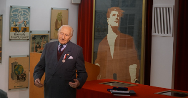 Senior Mikołaj Pac Pomarnacki stoi w muzuem Poznańskiego Czerwca 1956 na tle ekspozycji. Mężczyzna ubrany jest w garnitur, na piersi ma medal.