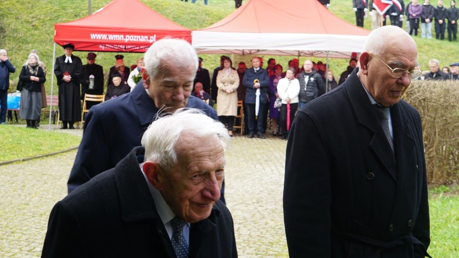 Na pierwszym planie trzech starszych mężczyzn ubranych w garnitury i czarne płaszcze. Mężczyzna idący z przodu niesie znicz. Na drugim planie ludzie stoją pod namiotami. W tle ściany fortu.