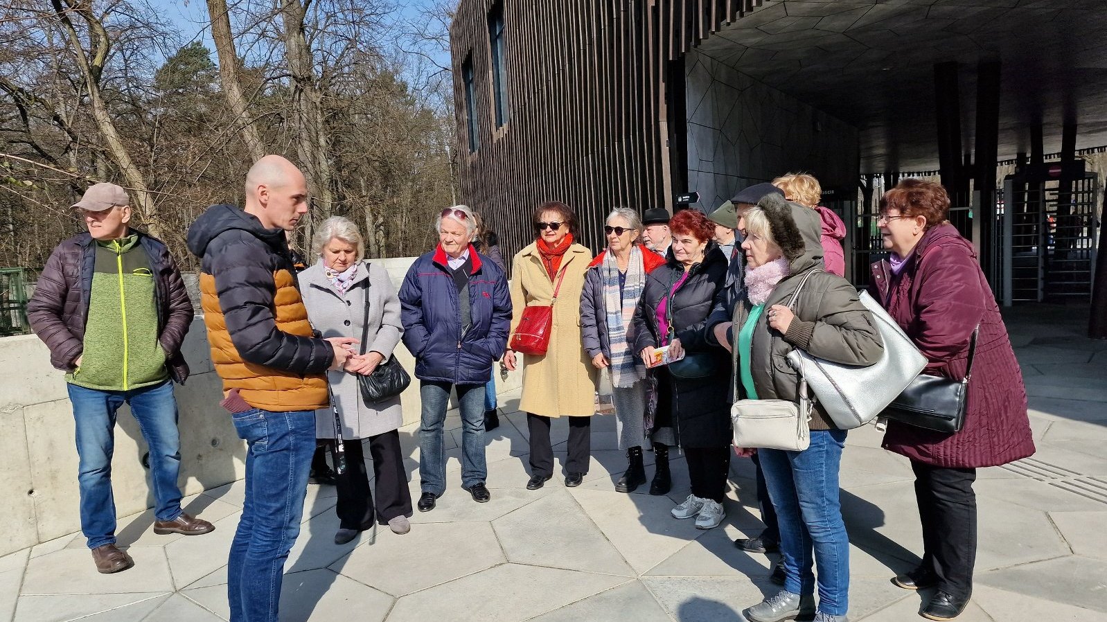 Zdjęcie przedstawia seniorów stojących przed wejściem do ZOO w Łodzi. Na zdjęciu przewodnik, w którego wpatrzeni są seniorzy.