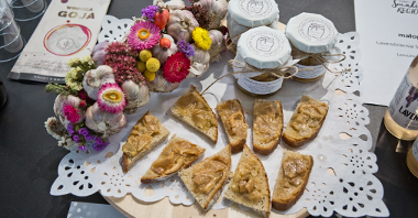 Na zdjęciu deska z pokrojonym chlebem, słoiczki z jedzeniem oraz kwiaty