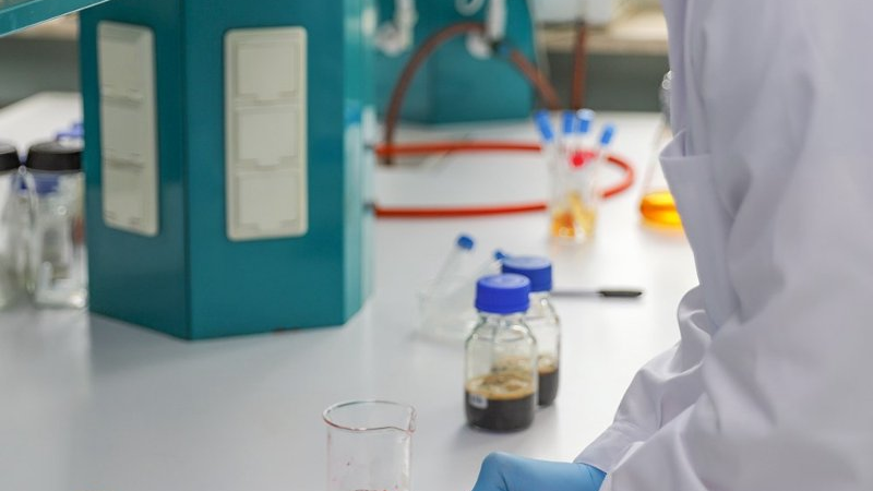 Zdjęcie, blat laboratorim, na którym stoją akcesoria badacza, nad blatem pochyla się laborant.