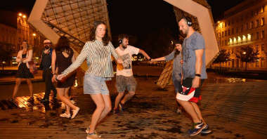 Zdjęcie przedstawia tańczących ludzi na placu Wolności. Za nimi widać fontannę.