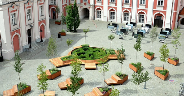Zdjęcie przedstawia dziedziniec urzędu miasta. Widać na nim budynek, ławki i drzewa w donicach.
