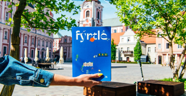 Na zdjęciu plac Kolegiacki, na pierwszym planie dłoń trzymająca książkę z napisem Fyrtle