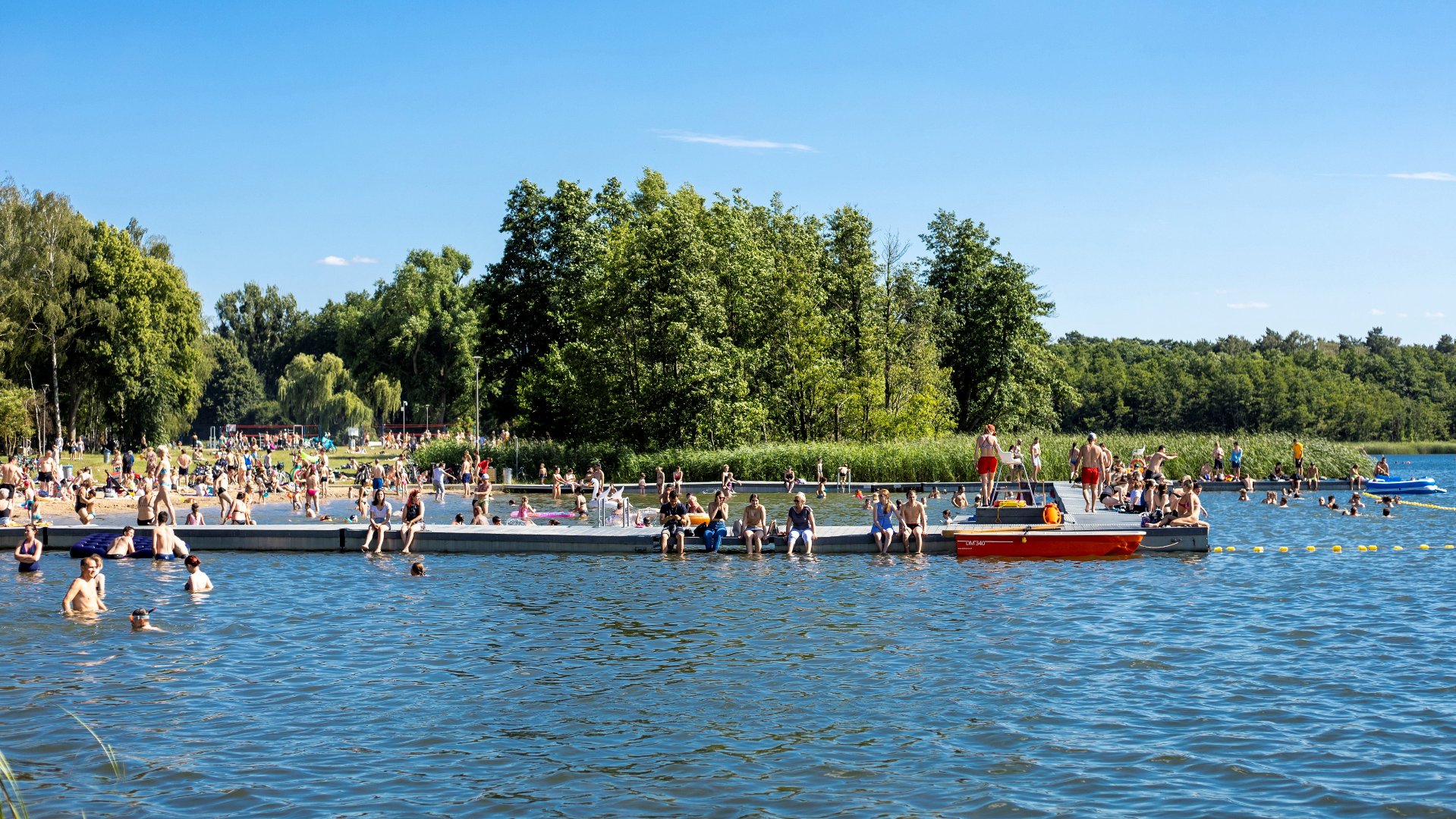 Ludzie siedzący na molo na poznańskim Jeziorze Strzeszyńskim. Po prawej stronie woda. Po lewej ludzie kąpiący się w wodzie i widok na ludzi na plaży. - grafika artykułu
