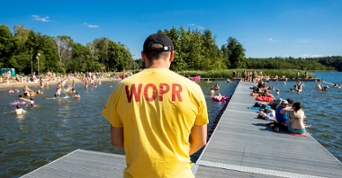 Grupa ludzi kapiących się w jeziorze. W centrum zdjęcia ratownik w żółtej koszulce z napisem WOPR.