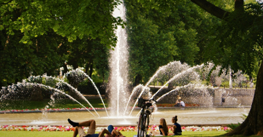 Grupę ludzi wypoczywających w cieniu drzew, w tle fontanna.