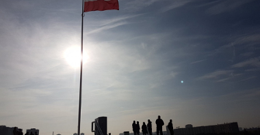 Popołudnie. Na szczycie Kopca stoi wysoki maszt z flagą Polski. Pod masztem stoją ludzie i oglądają widoki. W tle panorama Poznania.