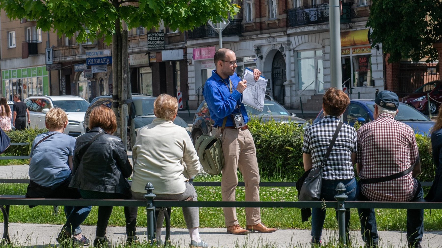 Przewodnik stoi do uczestników przodem i pokazuje mapę wycieczki. Opowiada uczestnikom o miejscu, w którym się znajdują. Uczestnicy siedzą na ławkach. W tle stare kamienice i zaparkowane samochody.