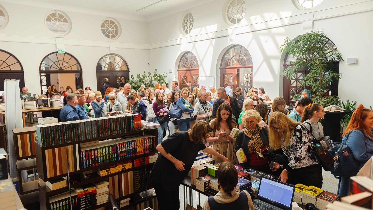 W oświetlonej sali stoją stoiska z książkami. Wokół stoisk ustawia się kolejka kupujących.