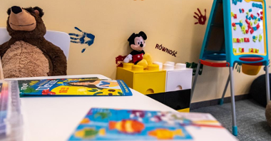 Na zdjęciu sala zabaw dla dzieci, na pierwszym planie stół z kolorowankami, w tle maskotki