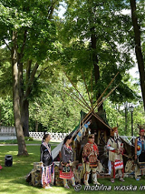 Indianie w charakterystycznych dla ich kultury strojach oraz namioty tipi. W tle trawa, drzewa i biały płot.
