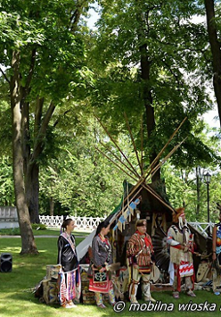 Indianie w charakterystycznych dla ich kultury strojach oraz namioty tipi. W tle trawa, drzewa i biały płot.