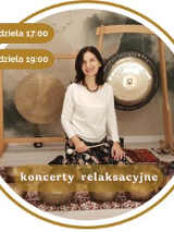 Kobieta w średnim wieku z czarnymi włosami. Ubrana w biały długi rękaw i wzorzyste spodnie. Siedzi na podłodze pokrytej dywanami. Za nią wiszą 2 gongi. Przed nią leży kilka mis.