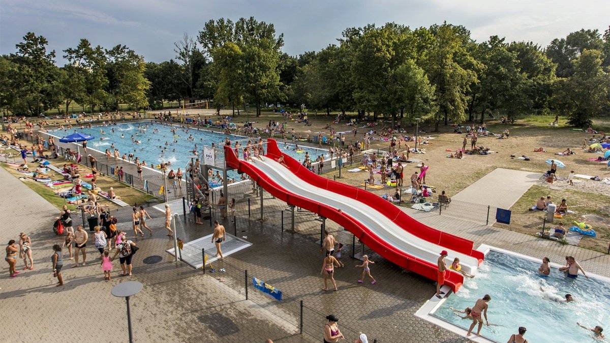 Dwa baseny letnie w Parku Kasprowicza. Między nimi zjeżdżalnia. Dookoła ludzie na ręcznikach. W basenach dorośli i dzieci. W tle drzewa.