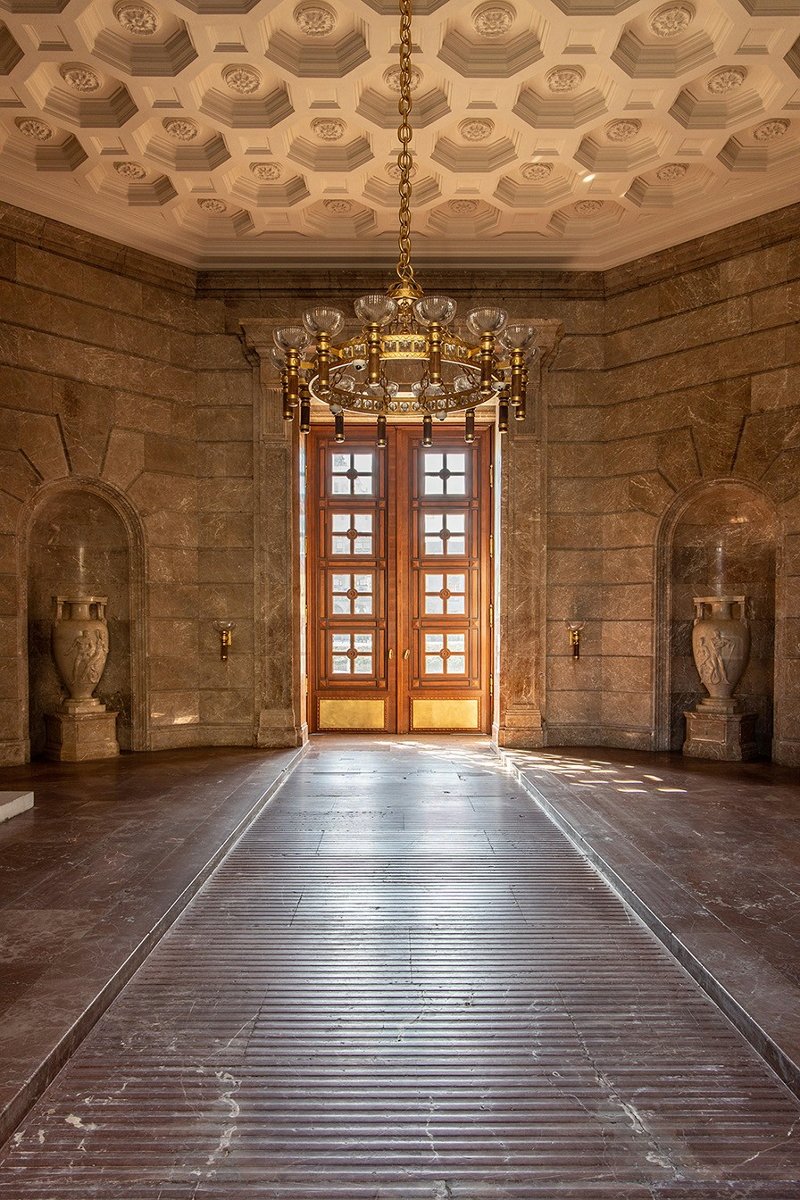 Zdjęcie z Sieni Przejazdowej z widokiem na duże, zabytkowe drzwi wejściowe. Drzwi zostały wykonane z drewna, wkomponowano w nie osiem kwadratowych okien. W centralnej części Sieni wisi historyczny żyrandol, a po bokach widoczne są duże wazy z marmuru.