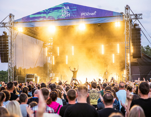Przed nami ostatnie koncerty z cyklu #NaFalach Publiczność stoi i pod sceną i bawi się do muzyki. Na scenie stoi mężczyzna z mikrofonem w ręce i unosi ręce do góry. Scena jest oświetlona na żółto. W tle drzewa.