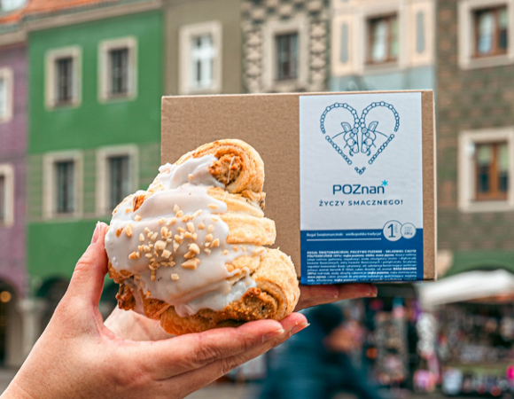 Rogal świętomarciński na tle kamieniczek na Starym Rynku. Za rogalem kartonik a na nim napis Poznań życzy smacznego.