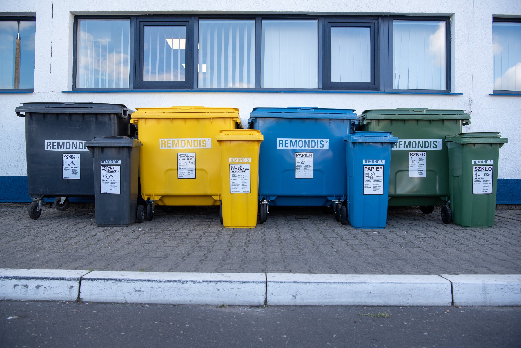Rząd czarnych, żółtych, niebieskich i zielonych dużych i małych kontenerów na śmieci, ustawionych przed białą fasadą budynku z oknami w ciemnych ramach.