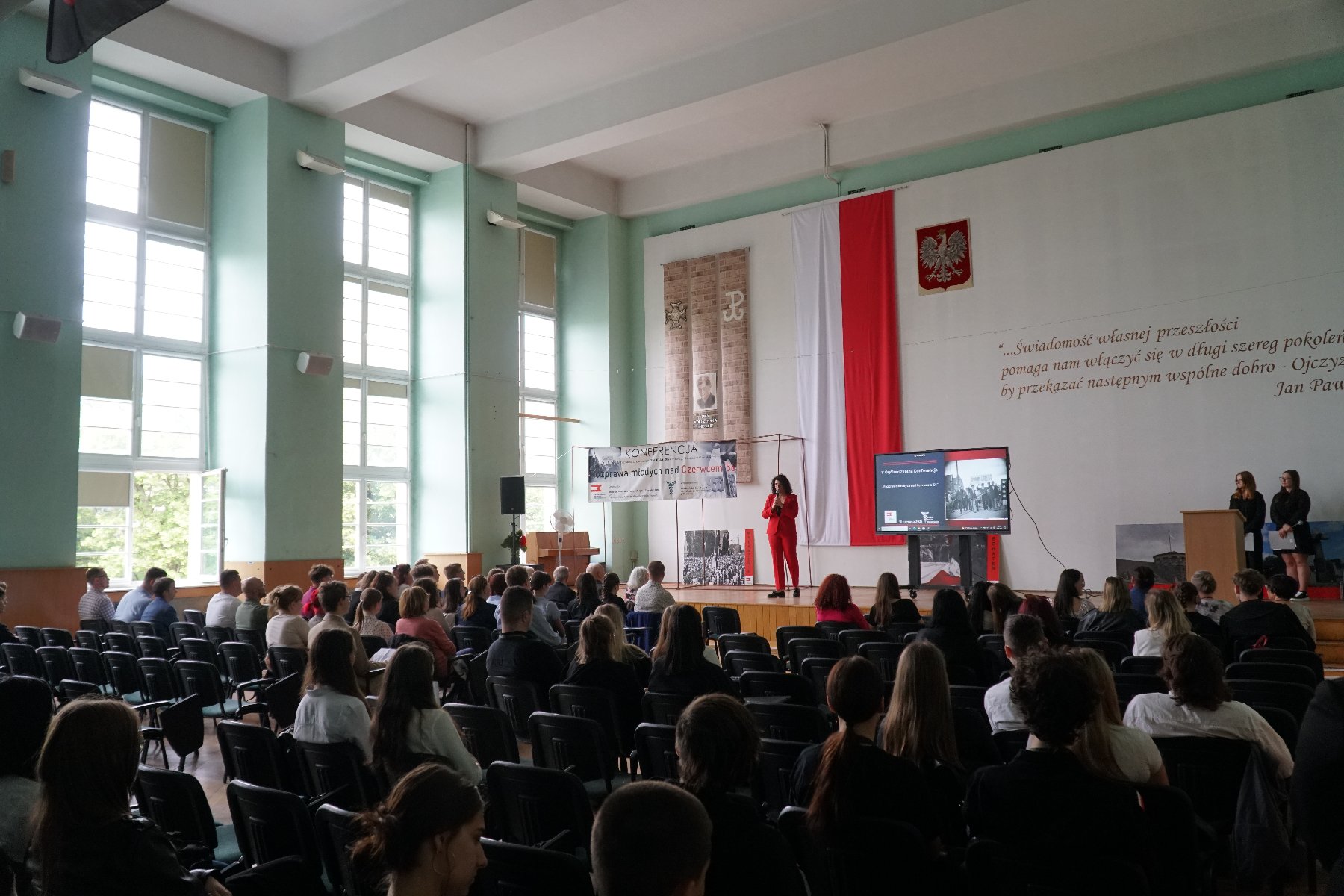 10 czerwca odbyła się V Ogólnoszkolna Konferencja "Rozprawa Młodych nad Czerwcem '56" Sala konferencyjna z dużymi oknami. Publiczność siedzi na krzesłach. Na scenie stoi kobieta w czerwonym garniturze, przemawia do uczestników. Za nią na ścianie duża flaga Polski, godło oraz cytat Jana Pawła II. Obok ekran z prezentacją. Po lewej baner z napisem "Konferencja - Wyprawa młodych nad Czerwiec'