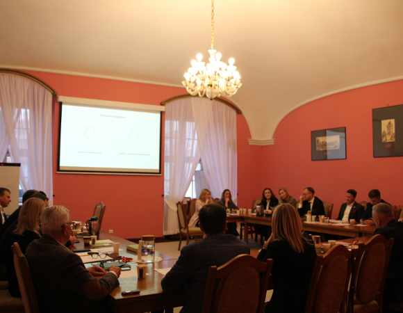 Sala Malinowa. Przy stole w kształcie podkowy siedzą kobiety i mężczyźni w eleganckich strojach i prowadzą rozmowy.