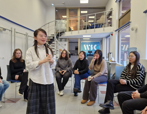 sala, na ustawionych w półokrąg krzesłach siedzą młodzi ludzie, którzy przysłuchują się temu co mówi stojąca w środku półokręgu młoda kobieta. Większość osób się uśmiecha