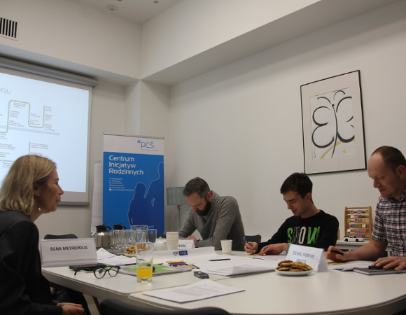 sala warszatowowa, przy stole siedzą cztery osoby. Pani Iwona Maruszczak-Szulc po jednej stronie stołu i trzech uczestników warsztatu po drugiej. Mężczyźni piszą coś na kartkach papieru. W tle na podświetlanej tablicy prezentacja z napisem "Mapa rozwoju".