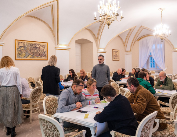 sala z ozdobnymi krzesłami, białe ściany ze złotymi zdobieniami. Przy stołach siedzą w grupach ludzie i pracują nad dokumentami