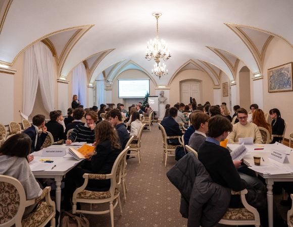 Sala z ozdobnymi krzesłami i złotymi zdobieniami na ścianach przy stolach siedzą grupki młodzieży pracującej w grupach