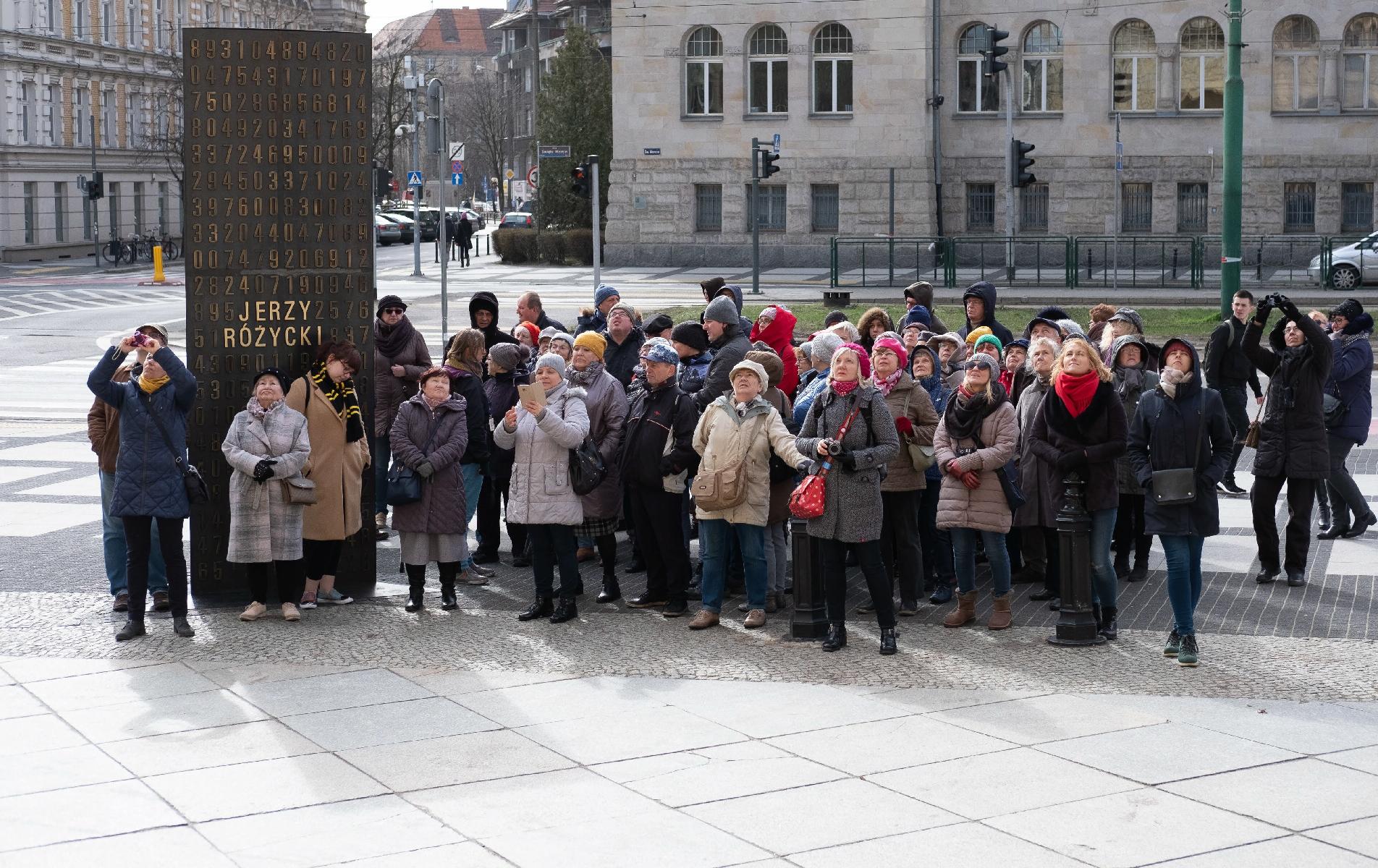 Grupa około 50 kobiet i mężczyzn stoi na ulicy Św. Marcin przy Pomniku kryptologów w Poznaniu. Zdjęcie zrobione podczas zimy. Ludzie ubrani są w płaszcze i kurtki, panuje chłodna atmosfera.