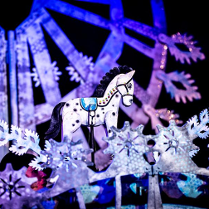 Zdjęcie ze spektaklu - biały drewniany konik wśród dekoracji w bialo-fioletowych kolorach.