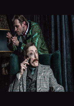 Zdjęcie z przedstawienia - jeden mężczyzna siedzący w fotelu z lupą przy oku, za nim stoi drugi mężczyna z fajką.