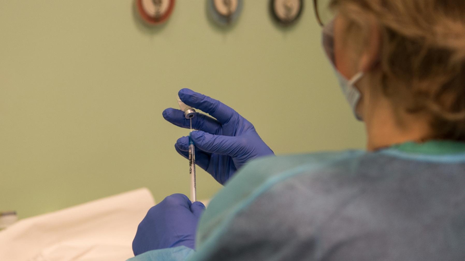 Stojąca tyłem kobieta w medycznym fartuchu, jedną dłonią (w rękawiczce) trzyma szczepionkę, drugą - nabiera ją strzykawką - grafika artykułu