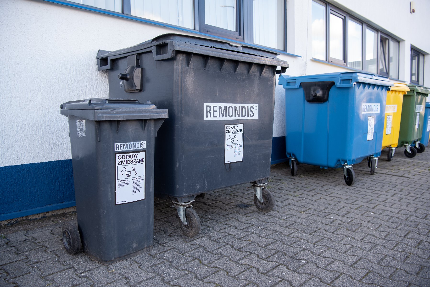 Szereg kolorowych kontenerów na śmieci firmy "REMONDIS" ustawiony wzdłuż białej ściany budynku na podłożu z kostki brukowej. Na pierwszym planie dwa ciemnoszare pojemniki z naklejkami "ODPADY ZMIESZANE", a dalej w rzędzie kontenery: niebieski, żółty oraz zielony.