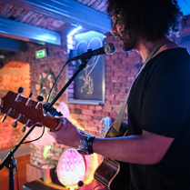 Mężczyzna z kręconymi włosami. Trzyma w rękach gitarę. Śpiewa do mikrofonu. W tle ceglana ściana z ozdobami.