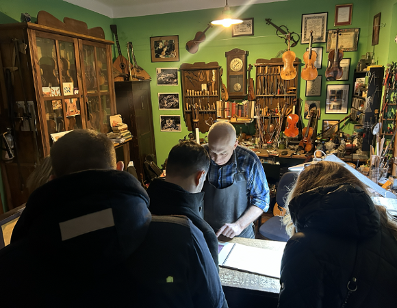 Uczestnicy wizyty studyjnej z Serbii przy stole, wspólnie oglądająca instrument muzyczny w pracowni lutniczej.