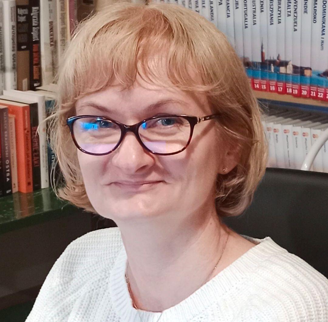 Anna Kleiber, fot. materialy organizatorów Uśmiechnięta kobieta w średnim wieku. Ma wlosy do uszu, grzywkę i okulary. Za nią regał z książkami.
