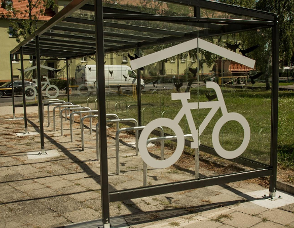 Wiata rowerowa z przeszklonym dachem i grafikami rowerowymi po bokach.